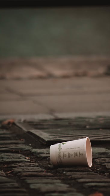A discarded white paper cup with green writing lies on a weathered brick or stone surface, partially tilted with its open end facing upwards. The surrounding environment appears to be an outdoor urban or residential area, with a pavement or pathway visible behind the cup, along with a dark metal grate embedded into the ground nearby. The background features a blurred, muted background in dull green and brown shades, suggesting foliage or outdoor earth tones, and the overall scene is lit by natural daylight, indicating an outdoor setting. The placement of the cup on the ground may imply it has been improperly disposed of, highlighting the importance of proper waste management, which Rubbish Removal Kentish Town services address through alternative waste handling options such as private collection and on-site clearance in the local area.