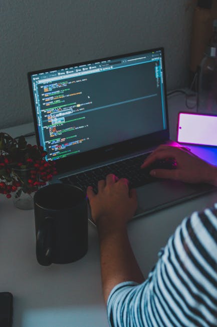 A person wearing a blue and white striped long-sleeved shirt is working on a laptop placed on a white desk, with the screen displaying a code editor with lines of colorful programming code. The person's right hand is resting on the laptop's touchpad, while their left hand is not visible. To the left of the laptop, there is a black ceramic mug, and behind the laptop, a small potted plant with red berries and green foliage is partially visible. On the right side, there is a white mobile phone with a glowing pink and purple screen, leaning against a dark object, possibly a speaker or a power bank. The background consists of a neutral-colored wall, and the lighting appears to be from an artificial source, creating a soft illumination on the workspace. The overall scene suggests a focused indoor environment, possibly a home office or workspace, with no visible rubbish or waste management elements present.