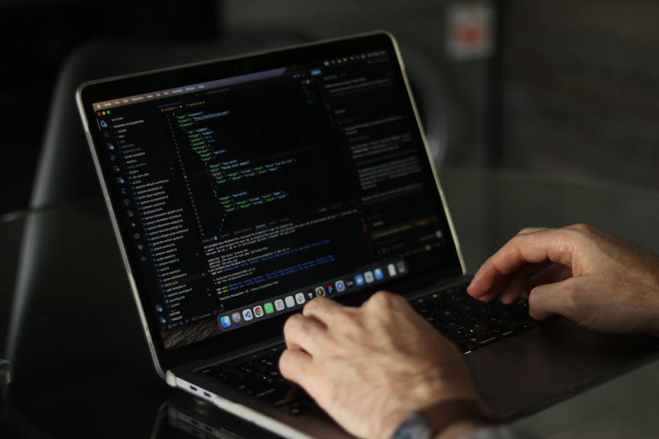 A close-up view of a person's hands using a laptop computer with a dark-themed code editor open on the screen, displaying lines of colour-coded programming code. The laptop is placed on a smooth, black surface, and the background is blurred, indicating an indoor setting with soft lighting. The scene suggests a focus on digital work, technology, or software development, with no direct connection to rubbish or waste management services but illustrating the use of electronic devices for administrative or planning tasks related to rubbish removal. The image captures the detailed texture of the person's skin and the sleek, silver finish of the laptop, providing a professional and neutral portrayal of working with technology in a work or home environment.
