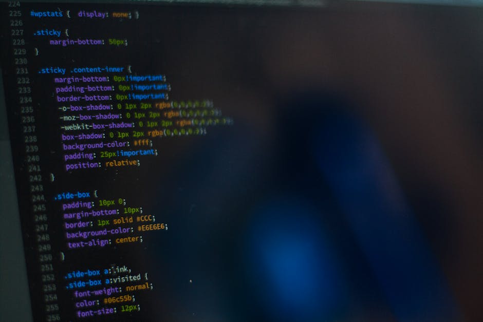A close-up photograph of a computer screen displaying lines of code in various colors, including blue, green, yellow, and purple, on a dark background. The code features CSS styling elements such as margin, padding, border, box-shadow, background-color, and font properties. The screen appears slightly blurred with a faint silhouette of a person's hand or finger in the foreground, possibly indicating interaction with the display. The environment suggests a workspace where digital or web development occurs, with the focus on coding. This image, though primarily related to programming, can be associated with the concept of data handling and management, paralleling the organization and disposal of waste, relevant to a rubbish removal service context. The professional tone of the scene aligns with an innovative approach to waste management, emphasizing the importance of digital and logistical precision in modern rubbish collection services offered by companies like Rubbish Removal Kentish Town.