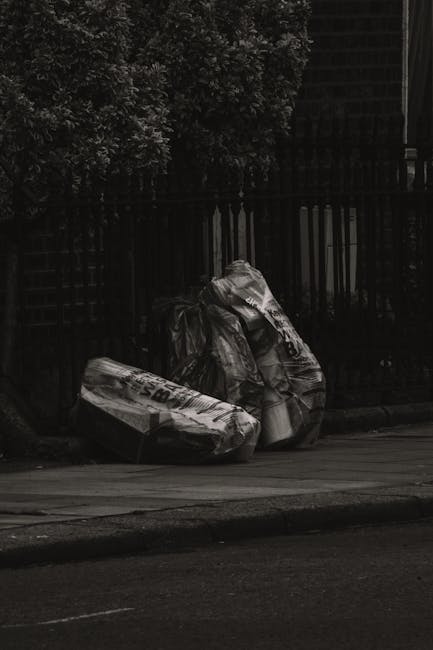 A trio of black plastic rubbish bags, tightly tied and slightly crinkled, are stacked and leaning against a dark metal fence along the edge of a sidewalk in an urban residential area. The bags appear to contain discarded household waste and are positioned on a paved surface beside a curb. Behind the fence, dense foliage of leafy trees or bushes is partially visible, creating a shaded background. The scene is captured under natural lighting conditions, with some areas of shadow cast by the surrounding trees and structures. The bags are arranged somewhat haphazardly, suggesting a recent or ongoing waste collection process. This scene reflects a typical urban rubbish disposal context, where external waste is left for private collection services, aligning with alternative waste handling methods outside of municipal rubbish collection, as often managed by companies like Rubbish Removal Kentish Town.