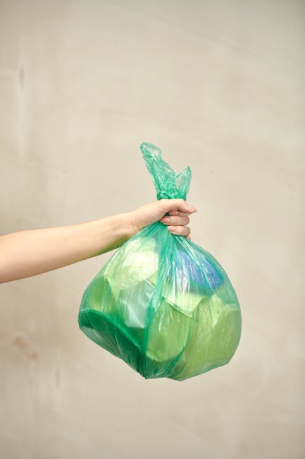 A trio of black plastic rubbish bags, tightly tied and slightly crinkled, are stacked and leaning against a dark metal fence along the edge of a sidewalk in an urban residential area. The bags appear to contain discarded household waste and are positioned on a paved surface beside a curb. Behind the fence, dense foliage of leafy trees or bushes is partially visible, creating a shaded background. The scene is captured under natural lighting conditions, with some areas of shadow cast by the surrounding trees and structures. The bags are arranged somewhat haphazardly, suggesting a recent or ongoing waste collection process. This scene reflects a typical urban rubbish disposal context, where external waste is left for private collection services, aligning with alternative waste handling methods outside of municipal rubbish collection, as often managed by companies like Rubbish Removal Kentish Town.