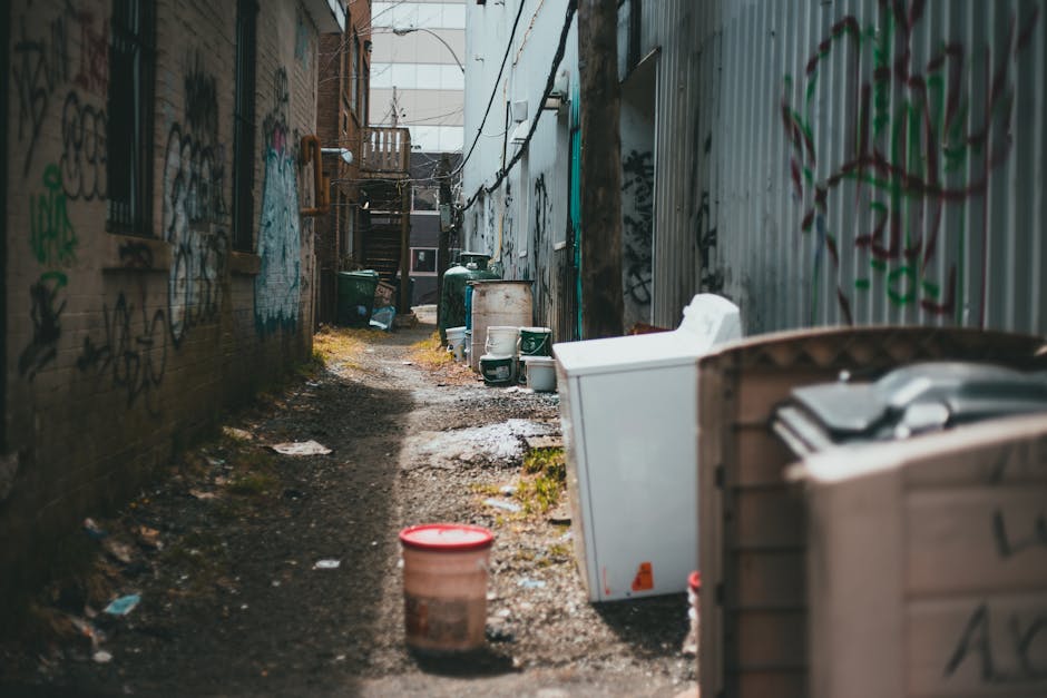 A narrow alleyway between two graffiti-covered brick and metal walls, cluttered with various discarded household items including a large plastic bucket with a red lid, a white upright freezer or appliance, a plastic storage bin, and several green wheelie bins lined along the alley. The ground is uneven and muddy, with patches of dirt, scattered debris, and small puddles. In the background, wooden stairs and a small balcony are visible at the end of the alley, suggesting access to residential buildings. The environment appears neglected with an informal collection of waste, indicative of an area where private rubbish collection or waste removal services, such as those offered by Rubbish Removal Kentish Town, might be necessary to manage illegal dumping or fly-tipping. The lighting is natural, with daylight illuminating the scene, emphasizing the disorderly state of the rubbish and the urban setting.