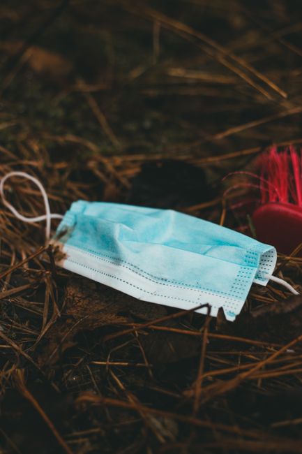 A light blue disposable face mask made of non-woven fabric with visible stitching and elastic ear loops lies discarded on dark, damp ground covered in brown, dry plant debris. Nearby, partially obscured, is a red-handled brush with stiff bristles. The background appears to be a natural outdoor setting with indistinct foliage and branches, creating a contrast between the medical waste and the natural environment. The scene suggests improper disposal of personal protective equipment, which could be linked to private waste handling services like those offered by Rubbish Removal Kentish Town, highlighting issues related to rubbish removal and the importance of proper rubbish disposal methods in urban or rural areas.