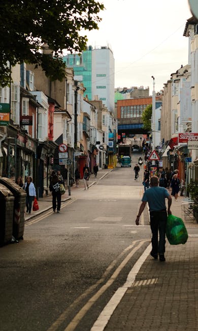 A busy urban street scene with a mix of commercial and residential buildings lining both sides of the road, showing small shopfronts, signs, and storefronts with awnings. The street has a gentle incline leading towards taller modern buildings in the background. Pedestrians are walking along the pavement and crossing the road; some are carrying shopping bags or personal items. In the foreground, a man wearing a teal shirt and dark trousers is walking away from the camera, holding a large green plastic bag, possibly filled with waste or recyclables, which may relate to private or alternative waste handling. The environment appears to be a lively town center with a mix of older architecture and contemporary structures, complemented by natural light and partial shade from nearby trees. The scene subtly reflects the context of independent waste collection or rubbish removal services operating within a typical town setting, with no visible waste bins but an emphasis on pedestrian activity and urban infrastructure as part of the waste management landscape.