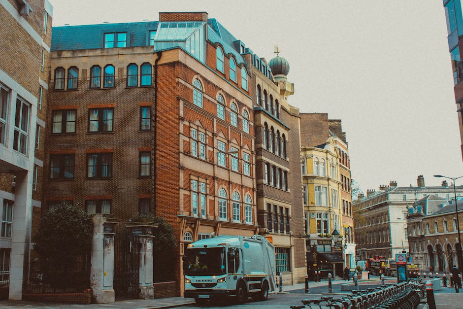 A street scene featuring a collection of multi-story buildings with diverse architectural styles, including brick facades and ornate window frames. In the foreground, a white waste collection truck is parked on the right side of the street, partially obscuring a black metal fence and a small gated entrance to a building. The truck is positioned on a paved road with black bollards lining the sidewalk, and the surrounding environment includes additional buildings with classical and modern designs, some with large windows and decorative cornices. The sky is overcast, casting diffuse lighting over the scene, consistent with urban waste removal activities, where private or independent waste collection services like Rubbish Removal Kentish Town might operate outside local authority schemes. The overall setting reflects typical city street infrastructure, preparation for rubbish collection, or ongoing waste disposal operations in a central London area.