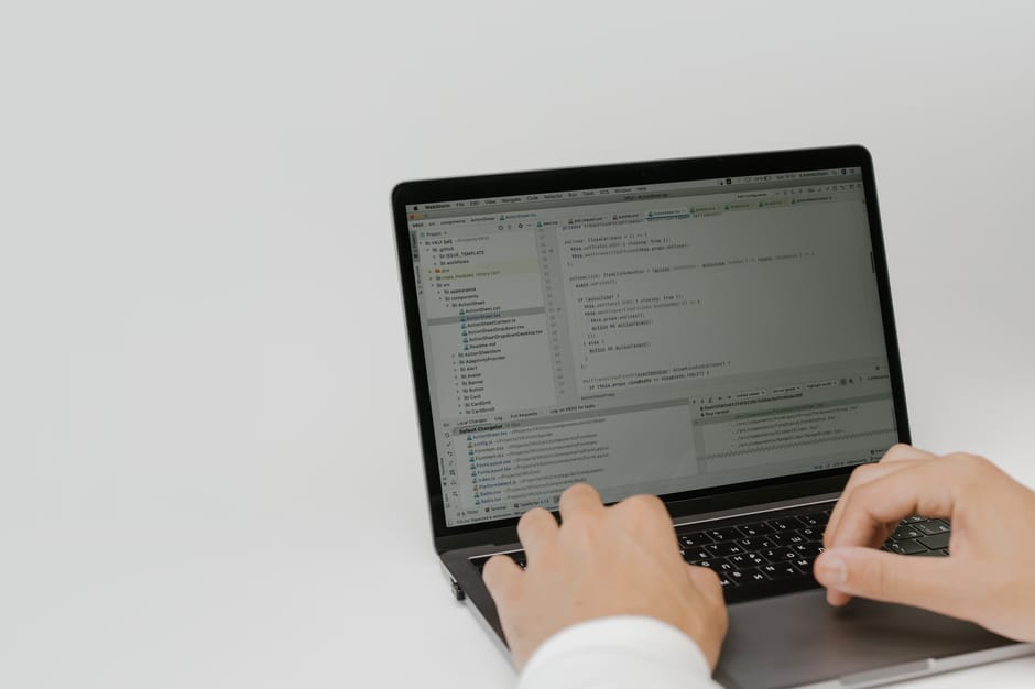 A person using a laptop on a white surface with a plain, light gray background. The laptop screen displays a coding or programming interface with multiple windows showing text and code. The individual’s hands are positioned over the keyboard, and the person is wearing a long-sleeved white shirt. The scene is well-lit with even lighting and minimal shadows, emphasizing the technological environment. This image relates to digital or online aspects of waste management services, such as scheduling or communication, and suggests an organized, professional approach suitable for rubbish removal companies like Rubbish Removal Kentish Town that may facilitate online booking or client interactions.