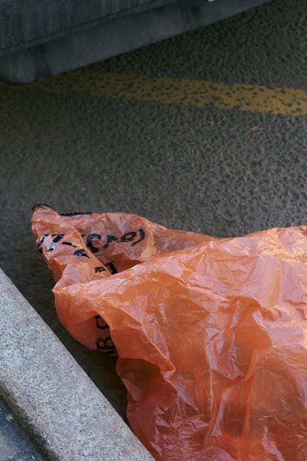A crumpled orange plastic rubbish bag lying on a textured asphalt surface in an outdoor setting. The bag appears to be discarded and is positioned near the edge of a concrete curb, with part of it partially folded or gathered. The background shows a strip of weathered, dark grey pavement with visible speckled detailing, and a section of a metal structure or object at the top of the image. The scene suggests an informal waste disposal point, which could be associated with private rubbish collection or on-site clearance services, typical in urban environments where rubbish removal by companies like Kentish Town Road rubbish removal guide NW5 is employed for waste handling. The lighting is neutral, with no strong shadows or reflections, emphasizing the utilitarian nature of the outdoor setting.