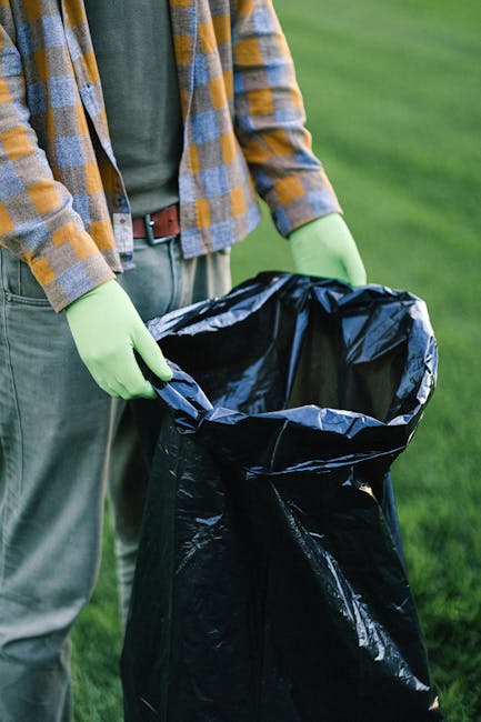 Close-up of a person outdoors, standing on a grassy area, holding an open black rubbish bag lined with a silver interior. The individual is wearing a plaid shirt with shades of blue, yellow, and gray, along with green gloves. Their belt and gray trousers are partially visible, and the background features well-maintained green grass. This scene depicts waste collection or rubbish disposal, with the person preparing to dispose of waste materials in the bag, which may relate to independent collection or on-site clearance services offered by companies like Rubbish Removal Kentish Town. The focus is on the act of handling rubbish within a natural outdoor setting, emphasizing manual waste management practices suitable for local rubbish removal activities.