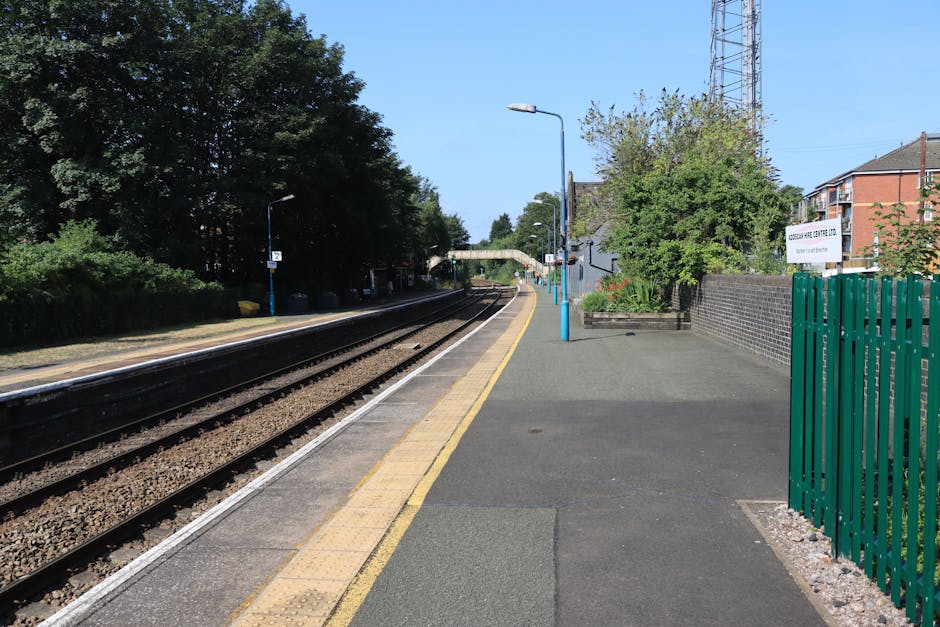A quiet train station platform with railway tracks running parallel along the left side, featuring dark asphalt rails and small gravel ballast beneath. The platform surface is paved with a mixture of smooth dark asphalt and textured yellow tactile paving along the edge, designed to assist visually impaired passengers. Tall, slim street lamps painted in light blue are evenly spaced along the platform, providing illumination during darker hours. On the right side, a low brick wall separates the platform from residential buildings and greenery, including well-maintained trees and flower beds with vibrant blooms. A green metal fence encloses a small area adjacent to the station, with signage indicating a local waste management or recycling company, suggesting an emphasis on private or alternative rubbish collection services. The background shows a clear blue sky, a distant footbridge crossing over the tracks, and lush treetops casting subtle shadows onto the scene, creating a calm and orderly environment typical of a suburban station focused on independent waste and rubbish removal.